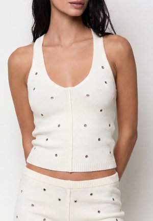 Donna che indossa un top bianco senza maniche in maglia e pantaloni abbinati decorati con piccoli occhielli metallici su uno sfondo semplice.