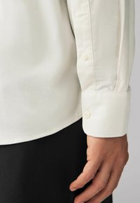 Chandail blanc en tissu côtelé avec un poignet boutonné. La main repose sur une surface noire, montrant une texture lisse et un design ajusté.