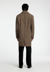 Cappotto marrone, con texture, colletto, maniche lunghe e taglio dritto, abbinato a pantaloni neri e scarpe, visto da dietro.