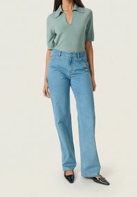 Vaalean siniset farkut suorilla lahkeilla, yhdistettynä vihreään lyhythihaiseen puseroon, jossa on V-pääntie ja kaulus. Mustat teräväkärkiset kengät.