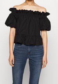 Schwarzes Oberteil mit schulterfreiem Design, Rüschenausschnitt und Puffärmeln, ausgestattet mit einem gestuften Saum und einem weichen, strukturierten Stoff. Kombiniert mit Jeans.