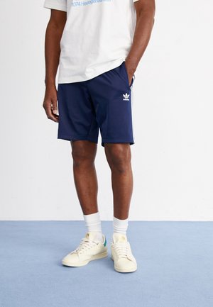 Homme portant un short Adidas bleu marine avec des bandes blanches, des chaussettes blanches et des baskets crème, debout sur un sol bleu clair.