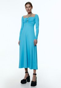 Langärmliges, figurbetontes Kleid in leuchtendem Blau, mit quadratischem Ausschnitt, gefälteltem Oberteil und einem Schlitzdetail an der Vorderseite. Schwarze Plateauschuhe.