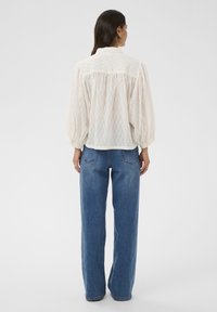 Blouse blanche avec motif texturé, manches bouffantes et col à boutons, associée à un jean bleu taille haute. Coupe simple et ample.