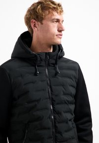 Zwarte ritsvest met een gewatteerde voorkant, fleece mouwen en een capuchon. Voorzien van verstelbare trekkoorden en een gestructureerde afwerking op de mouwen.