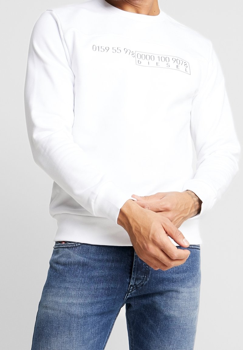 Bílá mikina s kulatým výstřihem, na přední části s raženým textem a čísly. Hladké manžety a lem; nošena přes modré džíny.