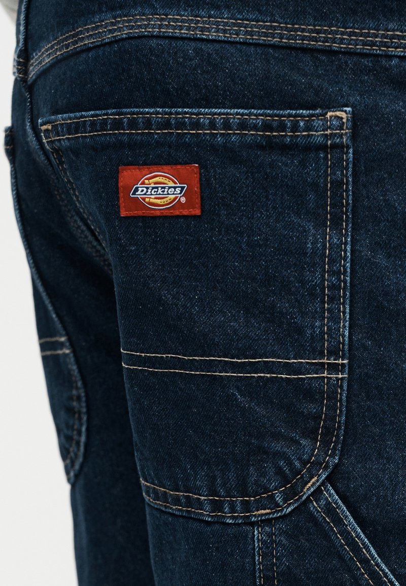 Tasca dei pantaloni in denim blu scuro con cuciture visibili e un'etichetta rettangolare rossa del marchio Dickies cucita nell'angolo in alto a sinistra.