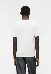 Λευκό βαμβακερό μπλουζάκι με κλασική στρογγυλή λαιμόκοψη και κοντά μανίκια, φορεμένο από άτομο που κοιτάζει προς τα πίσω, συνδυασμένο με σκούρο τζιν παντελόνι.