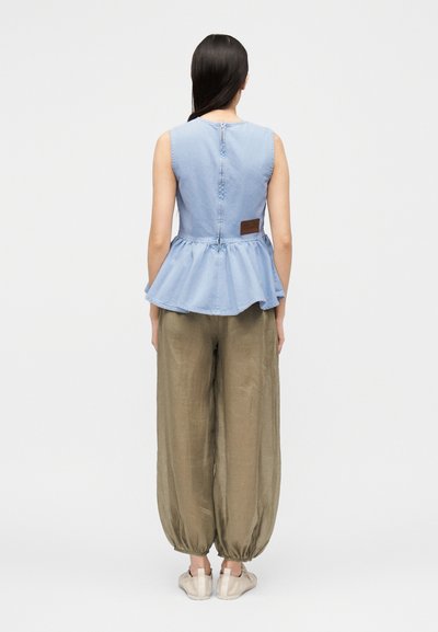 Femme debout, dos tourné, portant un top peplum bleu clair sans manches avec une fermeture éclair dans le dos et un pantalon ample vert olive serré aux chevilles.