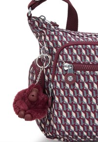 Schultertasche mit einem geometrischen Muster in Maroon und Creme. Mit einem plüschigen Affen-Anhänger und silbernen Hardware-Details. Sichtbare Reißverschlusstaschen.