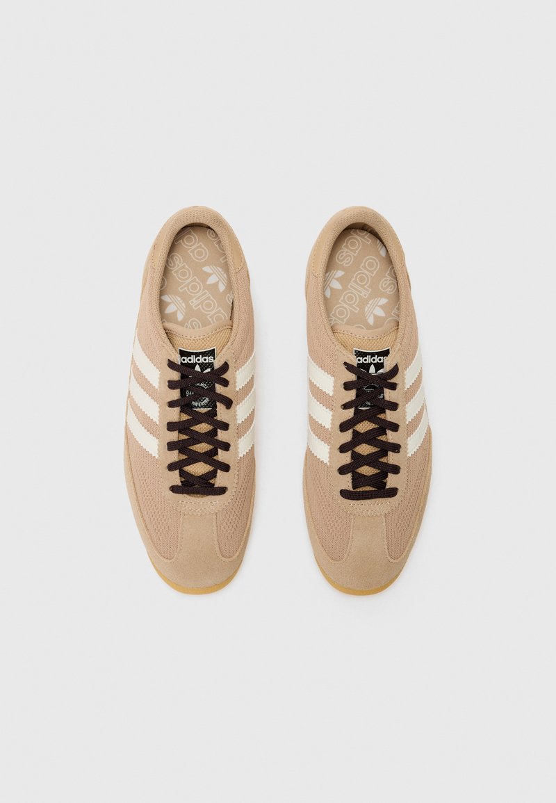 Zapatillas deportivas beiges con rayas blancas, material de ante y malla, suela de goma, cordones marrones y una etiqueta de logo en la lengua.