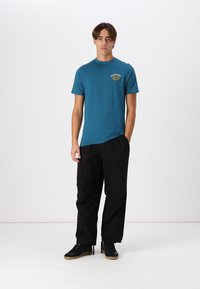 Teal cotton t-shirt with a front graphic logo, paired with black loose-fitting trousers and black sneakers. Smooth texture overall.