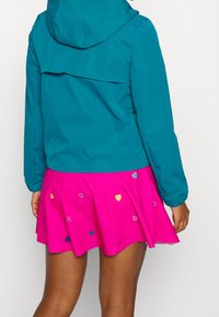 Turkos huvan jacka med elastiska ärmslut som matchas med en rosa utsvängd kjol med färgglada broderade former och en vågig fåll.