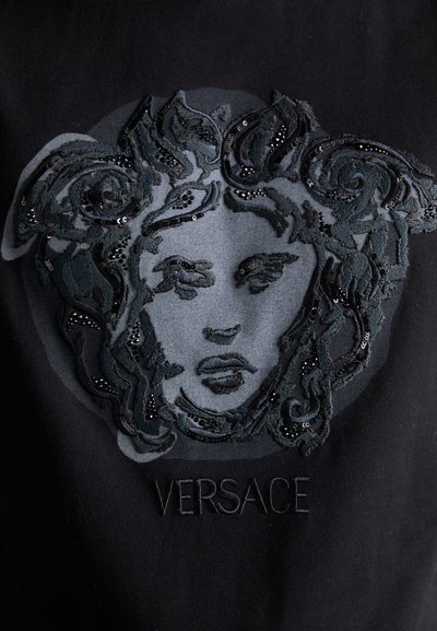 Black fabric featuring a textured, embroidered Medusa logo with sequins and intricate curly details, accompanied by the text "VERSACE" below.