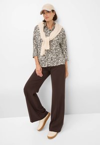 Cappello beige, blusa a pattern bianco e nero, maglione crema legato sulle spalle, pantaloni ampi marroni e sneakers beige con suola in gomma.