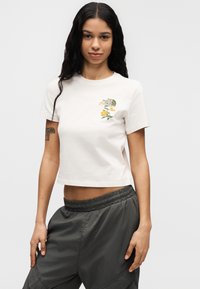 Λευκό κοντό t-shirt με κοντά μανίκια, διακοσμημένο με πράσινο και κίτρινο λουλουδένιο λογότυπο στο στήθος. Φορεμένο με γκρι παντελόνι με ελαστική μέση.