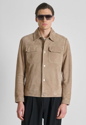 Man draagt een beige suède jas met klepzakken en een zwarte broek, kijkt recht vooruit, draagt donkere zonnebril, voor een effen lichte achtergrond.