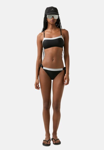 Černé bikiny s šedými akcenty, obsahující bandeau top a nízké kalhotky se bočními tkaničkami. Model má na sobě černou kšiltovku a oversized sluneční brýle.