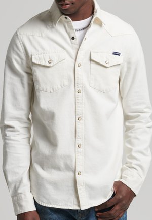 Mann trägt ein weißes Hemd mit zwei Brusttaschen, dunkle Jeans und eine silberne Kette vor einfarbigem Hintergrund.