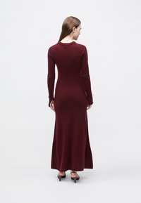 Bordowa dzianinowa długa sukienka z długimi rękawami, prążkowanym dekoltem i bocznym rozcięciem. Gładka faktura, dopasowana sylwetka.