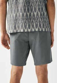 Grå shorts med en glat tekstur, der har en kantet kant. Toppen viser et indviklet mørkt og lyst geometrisk mønster.