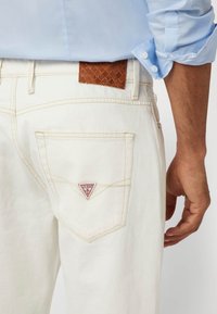 Balti džinsai iš baltos denim medžiagos su trikampiu raudonu logotipu ant užpakalinio kišenės. Virš juosmens esantis rudas tekstūruotas odinis ženklas turi prekės ženklo pavadinimą.