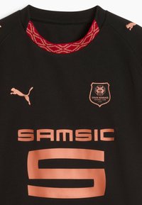 Melna futbola krekls ar īsām piedurknēm, sarkans rakstains apkaklītis, vara krāsas logo un teksts, kā arī izteikta Puma zīmola identifikācija uz pleciem.