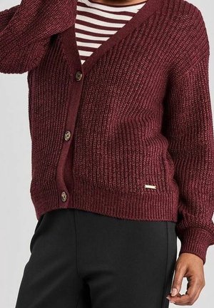 Frau trägt eine grob gestrickte, burgunderrote Strickjacke über einem gestreiften Hemd, kombiniert mit schwarzen Hosen, gezeigt von Hals bis Hüfte.