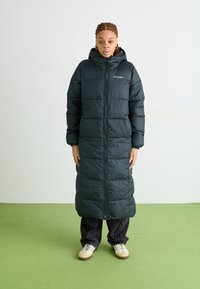 Tmavě zelená péřová bunda s kapucí, která má vodorovné prošívané části, plný zip a elastické manžety. Nosit se s volnými tmavými kalhotami.