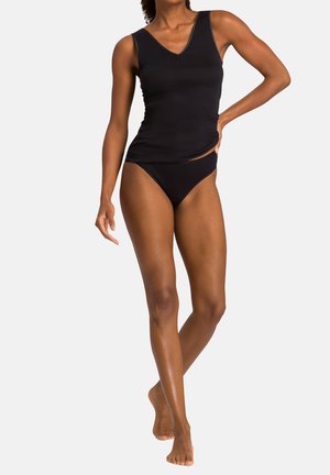 Donna che indossa una canotta nera senza maniche con scollo a V e slip bikini neri, in piedi con una mano sui fianchi e gambe incrociate.