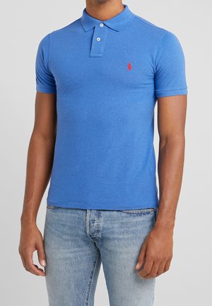 Homme portant un polo bleu ajusté à manches courtes avec un logo rouge sur la poitrine et un jean en denim délavé clair, sur un fond uni.