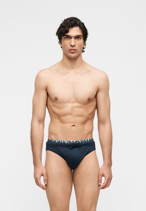 Homme musclé aux cheveux bouclés foncés portant un slip noir Emporio Armani, debout devant un fond gris clair uni.