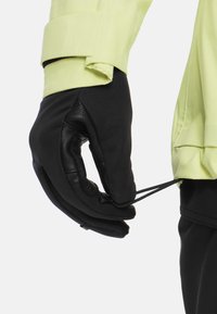 Schwarze Lederhandschuhe mit strukturiertem Griff, ausgestattet mit einem eng anliegenden schwarzen Stoffbund und einem limegrünen Jackenärmel, der eine Kordelzugdetails aufweist.