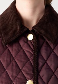 Gros plan sur une veste matelassée grenat avec un col en velours côtelé marron foncé et des boutons dorés fermés au niveau du cou.