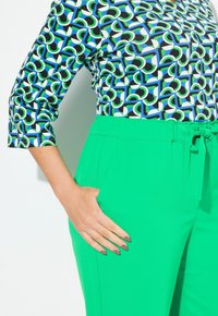 Zelené kalhoty s elastickým pasem a bočními kapsami v kombinaci s topem s vzorem, který obsahuje geometrické tvary v černé, zelené a modré barvě.
