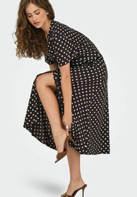 Robe midi noire à pois avec une coupe décontractée, des manches courtes et une fente sur le côté, associée à des sandales à talons hauts marron.