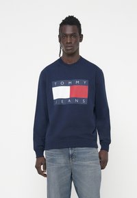 Felpa blu navy con scollatura a girocollo, caratterizzata da una grafica rettangolare bianca e rossa e testo "TOMMY JEANS" sulla parte anteriore.