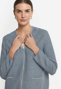 Hellblaue strukturierte Zip-Jacke mit zwei Fronttaschen und rundem Halsausschnitt. Das Material wirkt gewebt mit dezenten Musterdetails.
