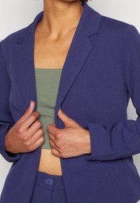 Marinblå blazer med en strukturerad design, skräddarsydd passform och slät textur, bärs över en grön åtsittande topp. Knapparna på byxorna är synliga.