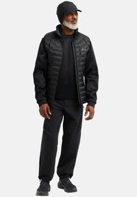 Gilet isolato nero sopra una camicia nera a maniche lunghe, abbinato a pantaloni cargo neri e scarpe leggere nere. Un cappellino nero completa il look.