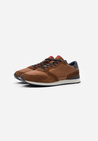 Brown suede sneakers with a smooth texture, dark blue laces, red detailing on the tongue, and a white midsole. Rubber sole provides grip.