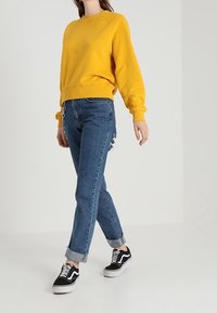Sudadera amarilla con cuello redondo, combinada con vaqueros azul claro con puños y zapatillas negras con suelas blancas.