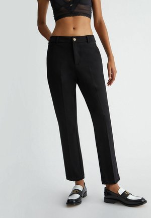 Zwarte, op maat gemaakte broek met rechte pijpen, voorzien van een knoopsluiting en strakke lijnen. Draagt bijpassende zwart-witte loafers voor een contrasterende look.