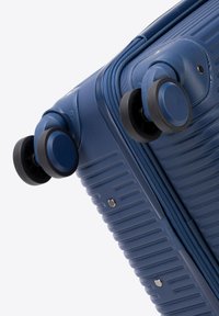 Valise rigide bleue avec une texture striée, quatre roues multidirectionnelles et une fermeture éclair. Les accents incluent des rivets en métal.