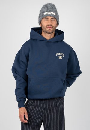 Man draagt een marineblauwe hoodie met de tekst "PROHIBITED", een grijze gebreide muts en donkere broek met krijtstrepen, staand tegen een lichtgrijze achtergrond.