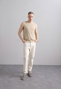Top sin mangas de canalé beige, jeans rectos blancos y zapatillas grises. Modelo de pie con las manos en los bolsillos frente a un fondo gris claro.