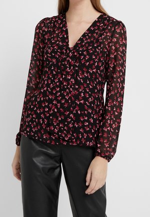 Femme portant un chemisier noir à motif floral rose avec de longues manches transparentes, associé à un pantalon en cuir noir.