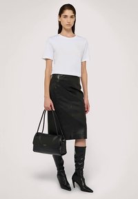 Falda de cuero negro con diseño ajustado, camiseta blanca de manga corta y botas negras de tacón hasta la rodilla. Se sostiene un bolso negro en una mano.