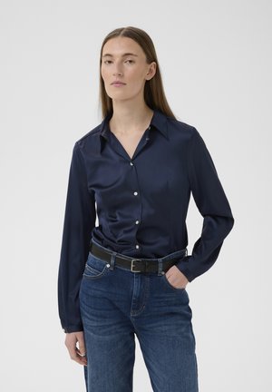 Žena s rovnými svetlohnedými vlasmi, oblečená v námorníckej saténovej košeli na gombíky, modrých džínsoch a čiernom opasku, stojaca s jednou rukou v vrecku.