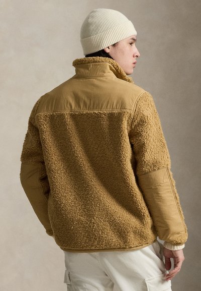 Φλις μπουφάν σε ταμπά απόχρωση με υφή βελούδου, αντίθετους λείους μανσέτες στους ώμους και μακριά μανίκια. Συνδυάζεται με λευκό ριμπ σκουφάκι.